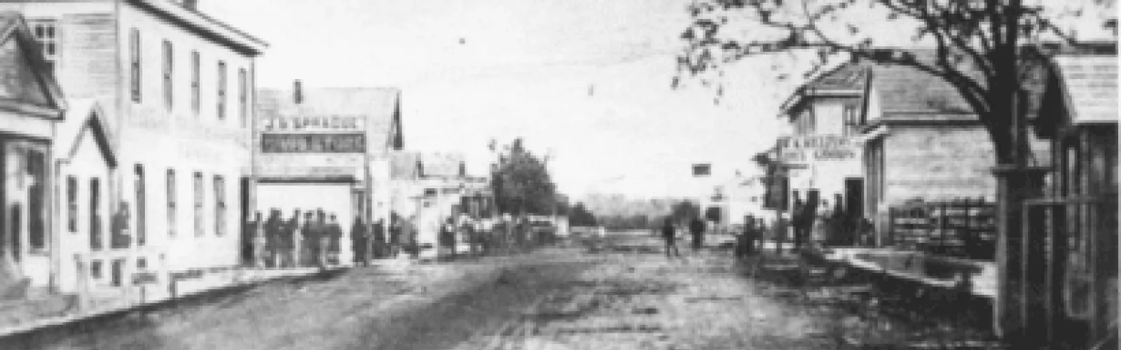 black and white photo of dirt road with buildings on each side with people on the sidewalks and street